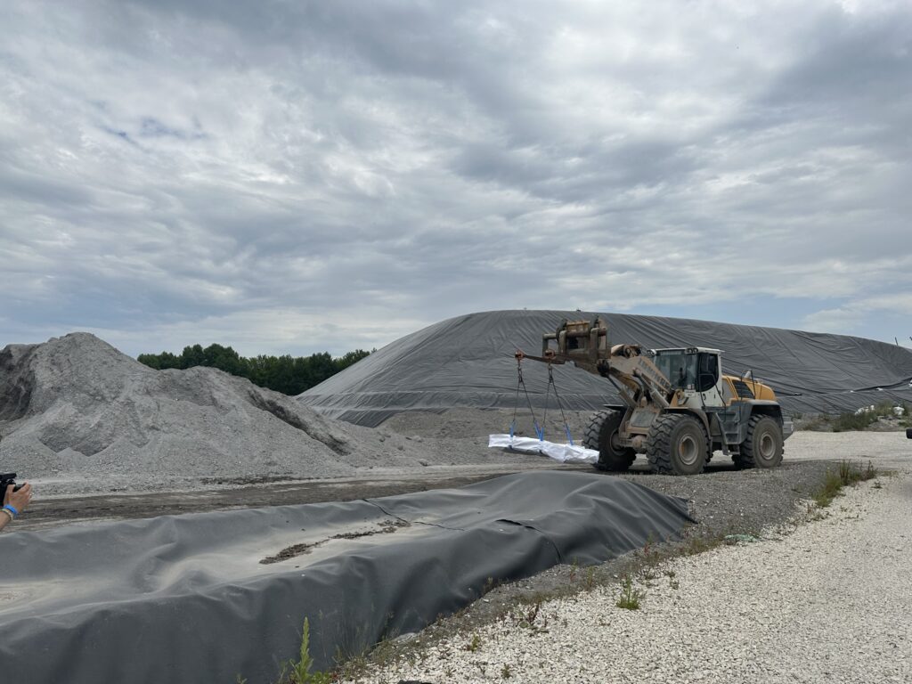 Ein Bagger hebt eine Last auf einer Baustelle mit grauen Schutthaufen und abgedecktem Material, unter bewölktem Himmel.
