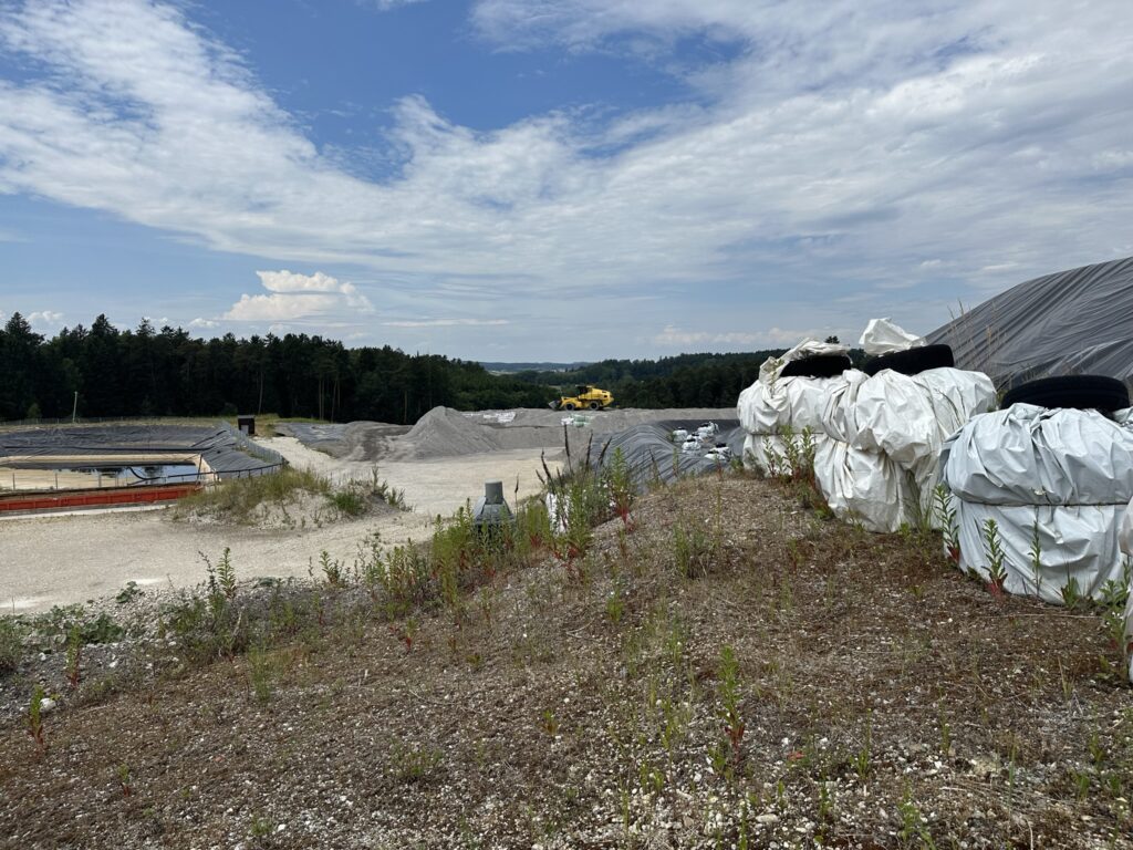 Bau- oder Industriegebiet mit gestapelten weißen Folienballen, Kieshügeln, Bagger im Hintergrund, bewölkter Himmel und bewaldete Landschaft im Hintergrund.
