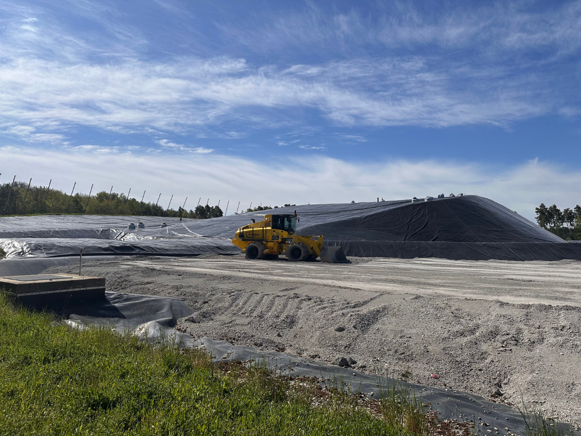 Ein gelber Radlader steht auf einer Baustelle neben einem großen, mit schwarzer Folie abgedeckten Erdwall unter blauem Himmel mit leichten Wolken.