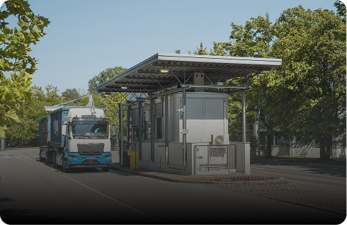 Ein Lastwagen fährt an einem modernen Kontrollposten mit Schranke vorbei, umgeben von Bäumen und klarem Himmel.