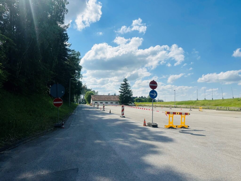 Straße mit Baustellenabsperrungen, mehreren Verbotsschildern und Wegweisern unter blauem Himmel mit Wolken, Wald links und Wiese rechts.