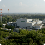 Großes Industriegebäude mit Schornsteinen, umgeben von Bäumen und Wald, unter einem leicht bewölkten Himmel.