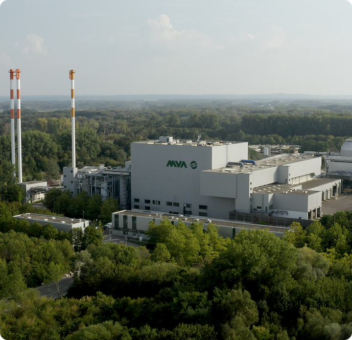 Große Industrieanlage mit weißen Gebäuden und zwei hohen Kaminen, umgeben von Bäumen und Wald, unter einem bewölkten Himmel.