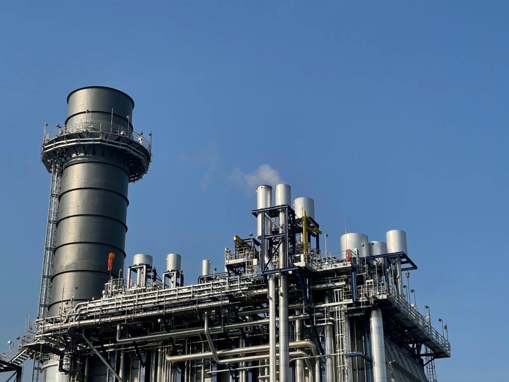 Industrieanlage mit großen Rohrleitungen und Schornsteinen unter klarem, blauem Himmel, etwas Rauch steigt auf.
