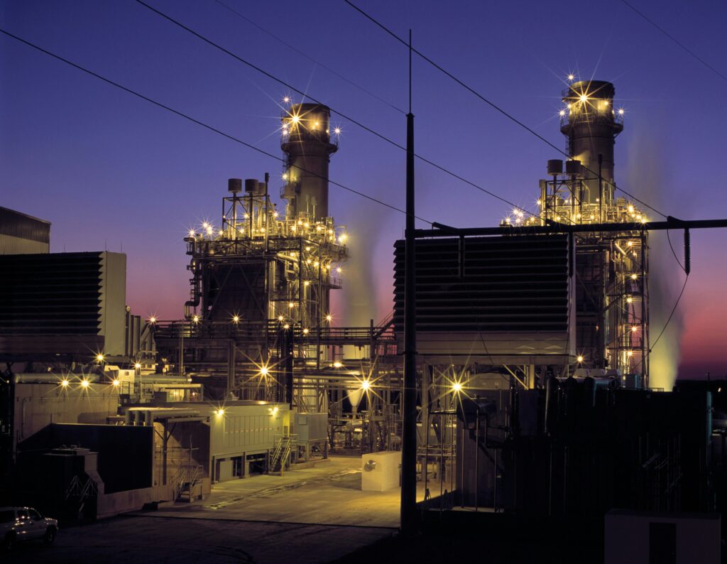 Ein Industriekomplex mit beleuchteten Türmen und Anlagen bei Nacht, viele Lichter leuchten, der Himmel ist dunkelblau.