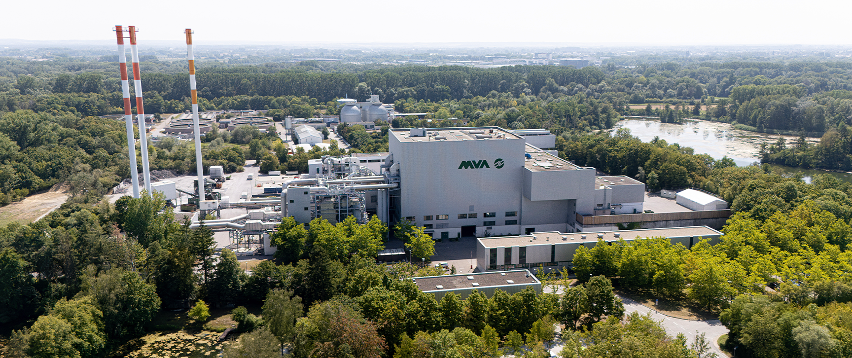 Großes Industriegebäude von AVA inmitten einer grünen, bewaldeten Umgebung, aufgenommen aus der Vogelperspektive an einem sonnigen Tag.