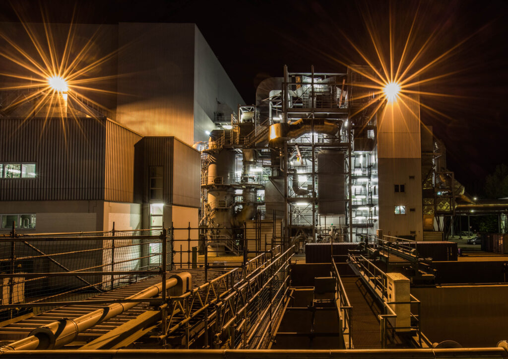 Industrieanlage bei Nacht, beleuchtet von gelbem Licht, mit sichtbaren Rohren und Strukturen im Vordergrund.