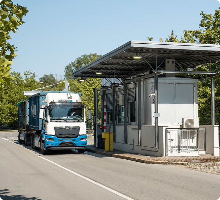 Ein Lastwagen fährt an einem modernen Kontrollpunkt mit Schranke vorbei, umgeben von Bäumen und blauem Himmel.