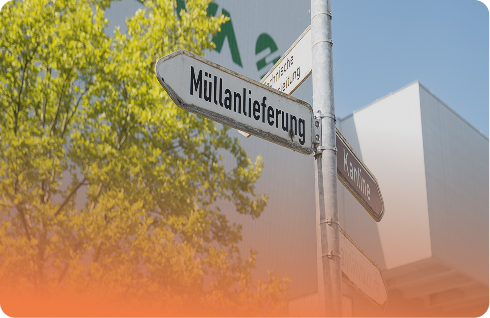 Straßenschild mit der Aufschrift "Müllanlieferung" vor einem Baum und einem Gebäude im Hintergrund.