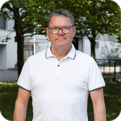 Ein älterer Mann mit Brille und weißem Polohemd steht draußen vor Bäumen und einem Gebäude.