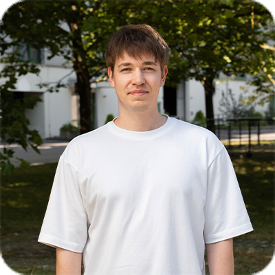 Ein junger Mann mit braunen Haaren steht im Freien vor Bäumen und Gebäuden und trägt ein weißes T-Shirt.