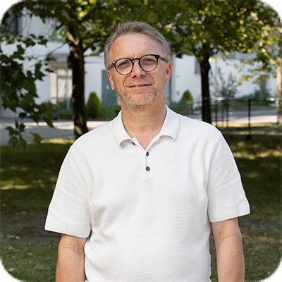 Ein älterer Mann mit Brille und weißem Poloshirt steht draußen vor Bäumen und einem Gebäude, lächelt leicht in die Kamera.