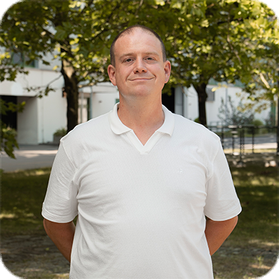 Ein Mann in weißem Poloshirt steht draußen vor Bäumen und einem Gebäude, schaut lächelnd in die Kamera.