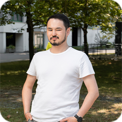 Ein Mann mit Bart steht draußen im Park, trägt ein weißes T-Shirt und posiert vor grünen Bäumen und Gebäuden im Hintergrund.