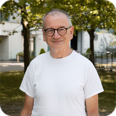 Ein älterer Mann mit Brille und weißem T-Shirt steht lächelnd draußen vor Bäumen und einem Gebäude.