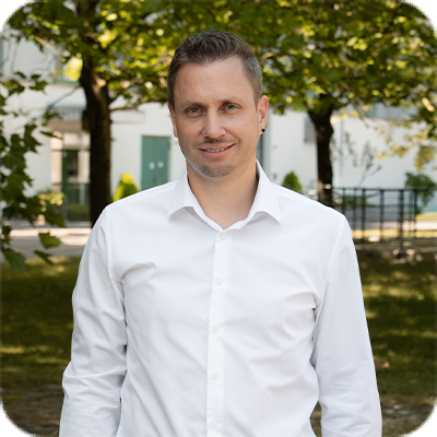 Mann mit kurzem Haar und weißem Hemd steht draußen vor Bäumen und einem Gebäude, lächelt in die Kamera.
