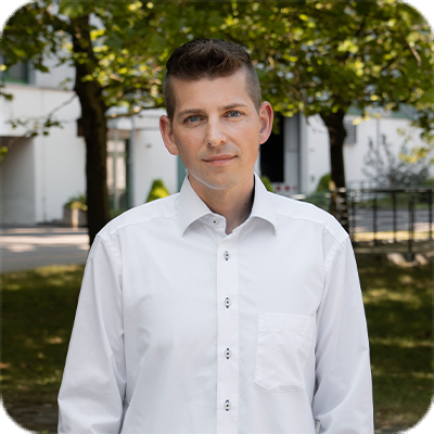 Ein Mann mit kurzem braunen Haar trägt ein weißes Hemd und steht draußen vor Bäumen und einem modernen Gebäude bei sonnigem Wetter.