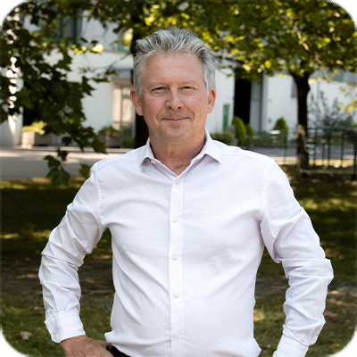 Ein älterer Mann mit grauen Haaren steht im Freien, trägt ein weißes Hemd und lächelt, Bäume und Gebäude im Hintergrund.