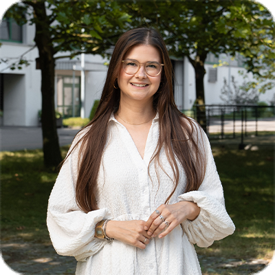 Eine Frau mit langen braunen Haaren und Brille steht draußen, trägt ein weißes Kleid und lächelt in die Kamera. Im Hintergrund sind Bäume und Gebäude zu sehen.