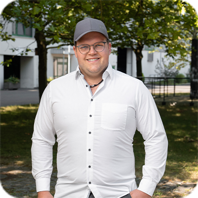 Ein Mann mit Brille und Mütze steht draußen, trägt ein weißes Hemd und lächelt. Im Hintergrund sind Bäume und Gebäude zu sehen.