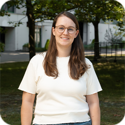 Frau mit langen braunen Haaren und Brille steht draußen vor einem Gebäude, trägt ein weißes T-Shirt und lächelt in die Kamera.