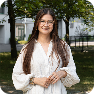 Frau mit langen braunen Haaren und Brille steht draußen, trägt ein weißes Kleid und lächelt, Bäume und Gebäude im Hintergrund.