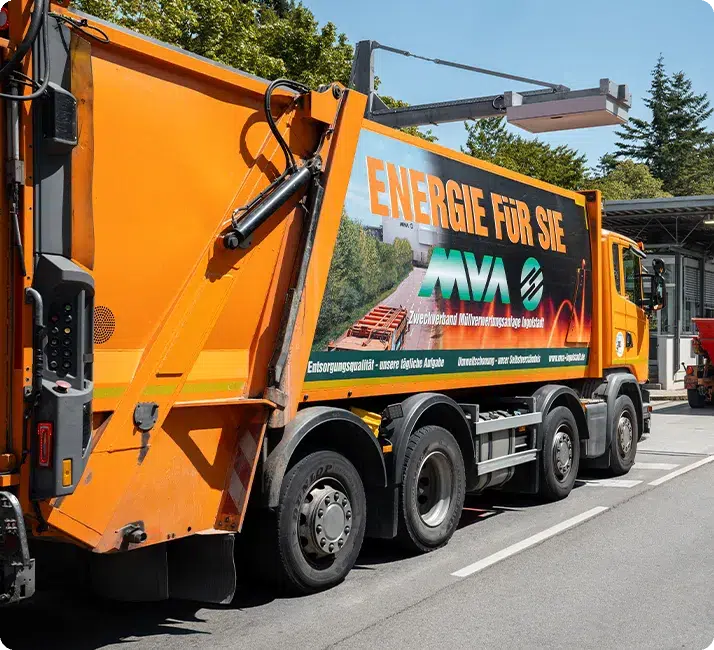 Oranger Müllwagen fährt auf einer Straße, Bäume im Hintergrund, Werbung mit Text "ENERGIE FÜR SIE" an der Seite des Fahrzeugs.