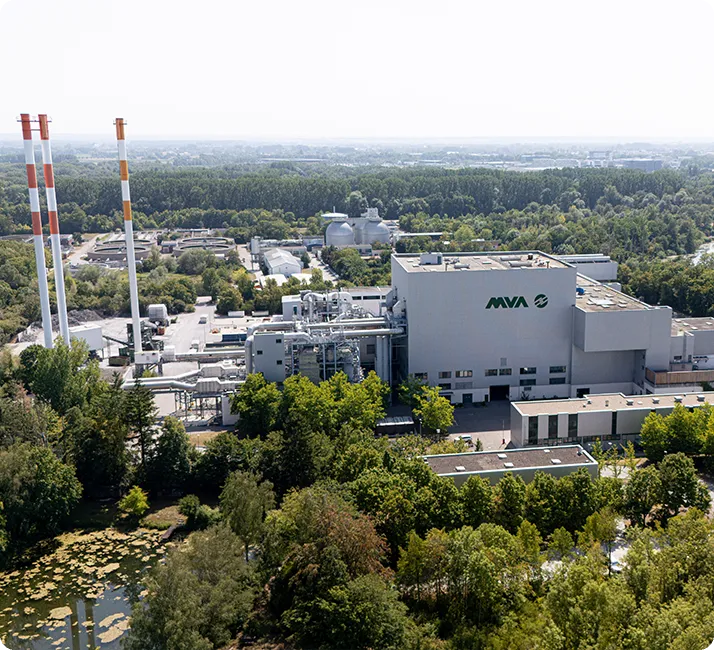 Industrieanlage mit großen Schornsteinen, umgeben von Bäumen und weiteren Gebäuden, im Hintergrund grüne Landschaft und bewaldete Flächen.