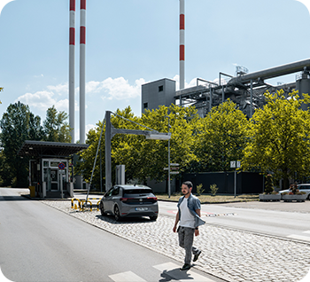 Ein Mann überquert eine Straße vor einem Industriegebäude mit zwei hohen Schornsteinen, einem geparkten Auto und grünen Bäumen im Hintergrund bei Tageslicht.