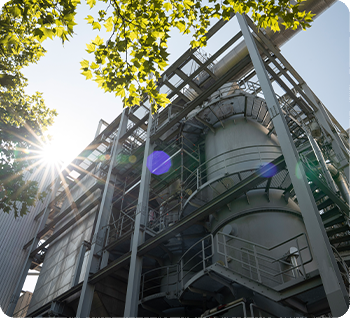 Industrielle Anlage mit Metallstrukturen und Rohren, Sonnenlicht scheint durch Bäume, modernes technisches Umfeld bei Tageslicht.