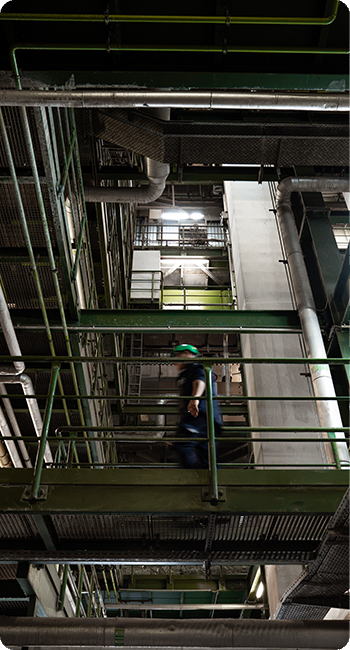Eine Person mit Schutzhelm geht durch eine industrielle Anlage mit Metallgeländern und Rohren. Die Umgebung wirkt technisch und industriell.