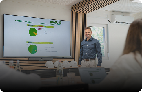 Ein Mann steht in einem modernen Konferenzraum vor einem Bildschirm mit Diagrammen und Präsentationsfolien, während mehrere Personen am Tisch sitzen.